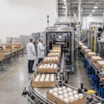packaging labor issues resolution - Korpack - Packaging Solutions for Complex Businesses Bottled products move along a conveyor on an automated packaging line in a clean factory, with two workers in white coats and hairnets overseeing the process.