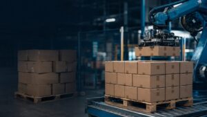 Your Best Packer and Your Worst Packer - Korpack - Packaging Solutions for Complex Businesses Industrial robotic arm lifts a cardboard box onto a pallet in a dimly lit warehouse environment
