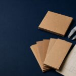 ChatGPT Image Jul 24 2026 11_53_57 AM - Korpack - Packaging Solutions for Complex Businesses Pile of brown cardboard sample cards with a metal caliper and a technical blueprint on a dark blue surface.