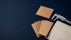 ChatGPT Image Jul 24 2026 11_53_57 AM - Korpack - Packaging Solutions for Complex Businesses Pile of brown cardboard sample cards with a metal caliper and a technical blueprint on a dark blue surface.