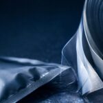Meat-Snack Packaging Is All About the Barrier - Korpack - Packaging Solutions for Complex Businesses Roll of clear plastic film with a sealed pouch on a dark surface, showing packaging materials ready for use.