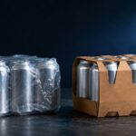bev multipack changes - Korpack - Packaging Solutions for Complex Businesses Two six-pack beverage cans: one in clear plastic wrap, the other in a brown cardboard carrier on a dark surface.
