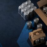 craft drink secondary packaging - Korpack - Packaging Solutions for Complex Businesses Assorted packaging supplies on a dark blue surface, including metal cans in boxes and a roll of packing tape (Korpack branding).
