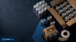 craft drink secondary packaging - Korpack - Packaging Solutions for Complex Businesses Assorted packaging supplies on a dark blue surface, including metal cans in boxes and a roll of packing tape (Korpack branding).