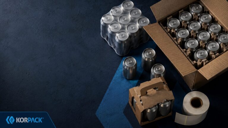 craft drink secondary packaging - Korpack - Packaging Solutions for Complex Businesses Assorted packaging supplies on a dark blue surface, including metal cans in boxes and a roll of packing tape (Korpack branding).