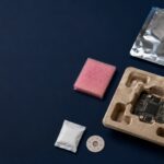 electronic packaging issues - Korpack - Packaging Solutions for Complex Businesses Circuit board partially removed from a molded pulp tray, with pink foam, a desiccant packet, a small white packet, a circular badge, and a silver anti-static bag on a dark blue background.