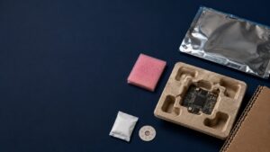 electronic packaging issues - Korpack - Packaging Solutions for Complex Businesses Circuit board partially removed from a molded pulp tray, with pink foam, a desiccant packet, a small white packet, a circular badge, and a silver anti-static bag on a dark blue background.