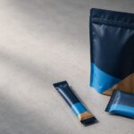 protein snack packaging playbook - Korpack - Packaging Solutions for Complex Businesses Blue and tan resealable snack pouch (standing) with two matching small wrappers on a gray concrete surface.