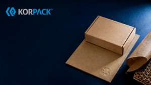 ChatGPT Image Aug 11 2026 01_53_01 PM - Korpack - Packaging Solutions for Complex Businesses KORPACK logo on a dark blue background with brown cardboard boxes and packing material nearby in a studio setup.