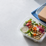 ChatGPT Image Aug 13 2026 03_25_53 PM - Korpack - Packaging Solutions for Complex Businesses Container of fresh salad with cherry tomatoes, cucumbers, chickpeas, grains, feta, and a creamy dressing in a small cup, on a blue napkin.