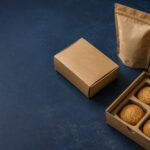ChatGPT Image Aug 6 2026 11_31_54 AM - Korpack - Packaging Solutions for Complex Businesses Kraft cardboard cookie box open with four cookies inside, plus a standing resealable pouch on a dark blue surface.
