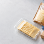 cheese packaging materials - Korpack - Packaging Solutions for Complex Businesses Assortment of cheese: sliced cheese in a clear tray, shredded cheese in a stand-up pouch, and a cheese sauce in a cardboard box on a blue cloth backdrop.