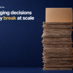 Scale Into National Retail - Korpack - Packaging Solutions for Complex Businesses Dark blue backdrop with a tall stack of flattened cardboard on the right; on the left, the headline reads 'SCALING INTO NATIONAL RETAIL' and 'The packaging decisions that quietly break at scale' (highlighted word 'break').