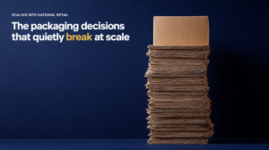 Scale Into National Retail - Korpack - Packaging Solutions for Complex Businesses Dark blue backdrop with a tall stack of flattened cardboard on the right; on the left, the headline reads 'SCALING INTO NATIONAL RETAIL' and 'The packaging decisions that quietly break at scale' (highlighted word 'break').